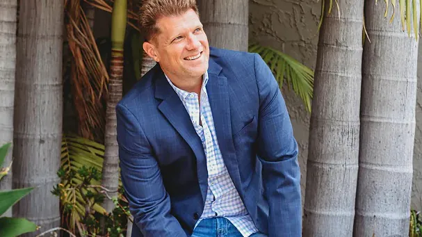 Smiling man in a blazer sitting among tropical plants, showcasing a relaxed and stylish outdoor vibe.